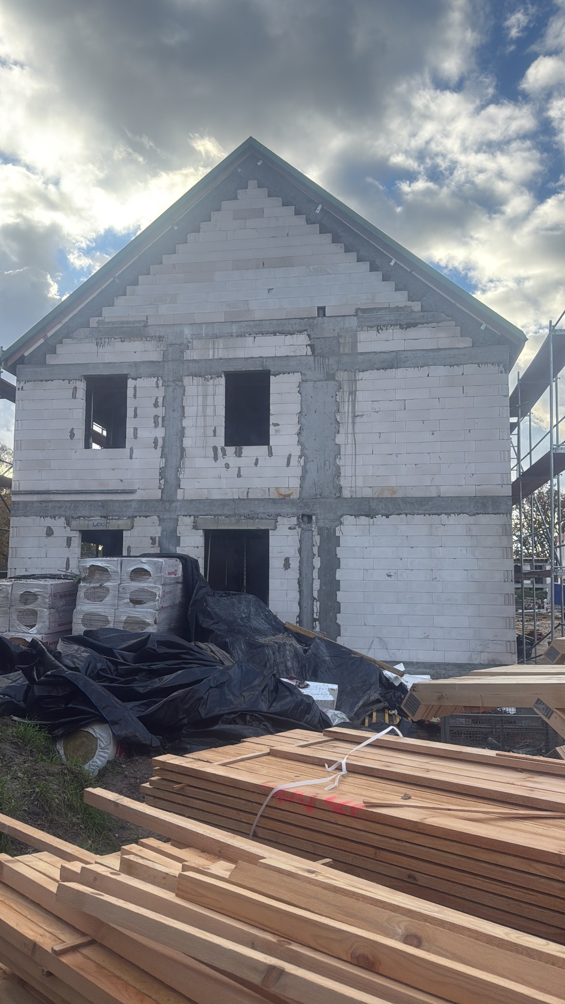 Wznoszenie dwukondygnacyjnego domu z białych bloczków, widoczne pakiety materiałów budowlanych i drewno na pierwszym planie, pochmurne niebo w tle.