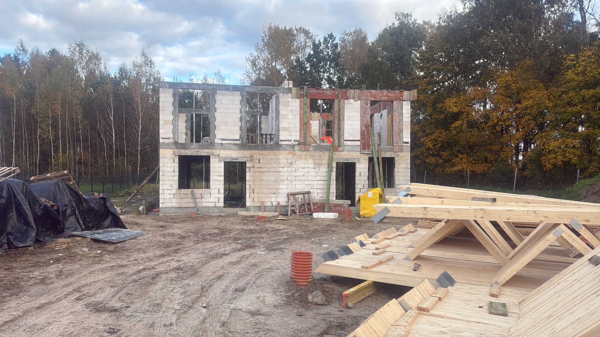 Dwukondygnacyjny budynek w trakcie murowania ścian z betonu komórkowego, widoczne drewniane elementy konstrukcyjne dachu i las. Jesienna sceneria, pochmurne niebo.