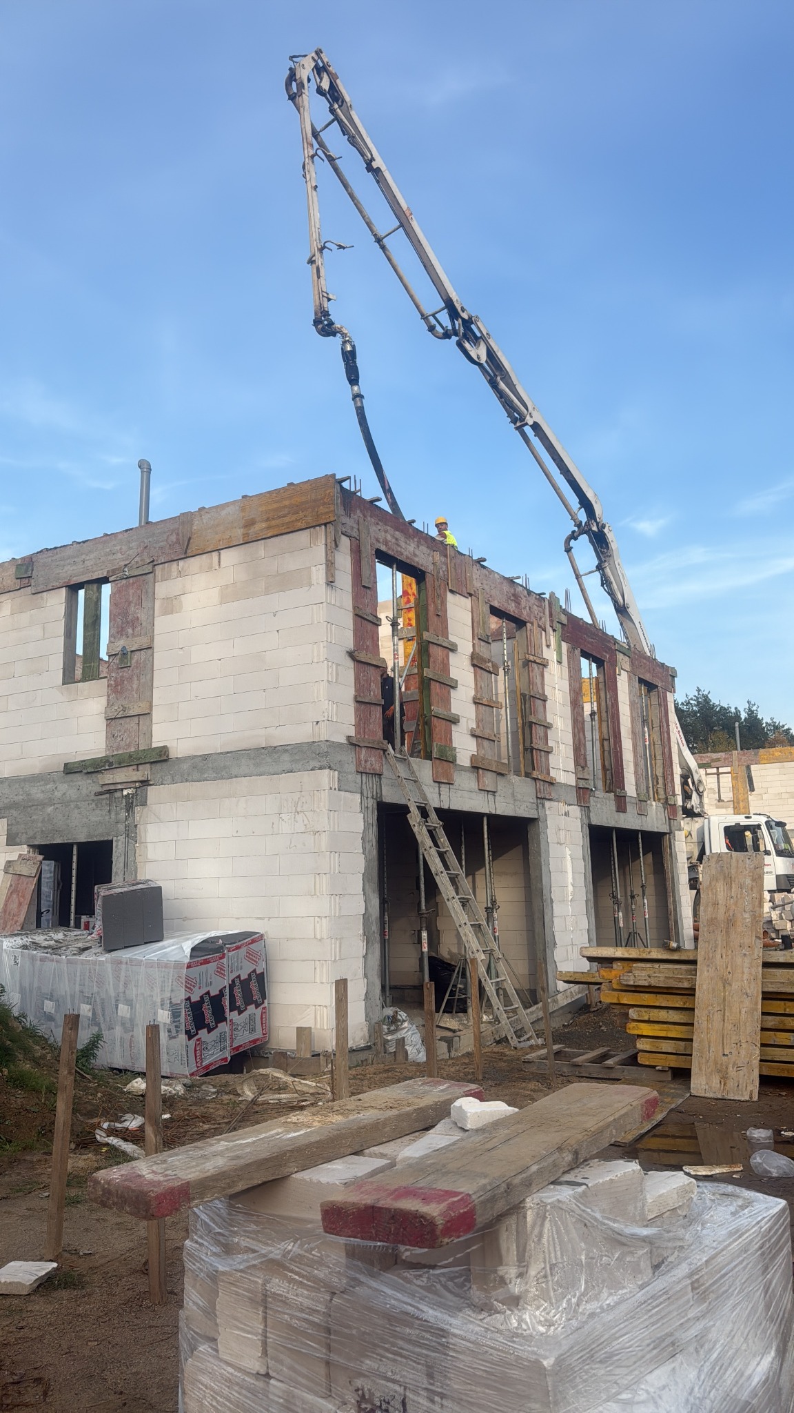 Wznoszenie dwukondygnacyjnego budynku z betonu komórkowego. Na górze pracownik w kasku obsługuje pompę do betonu. Widoczne szalunki okienne i stropowe.