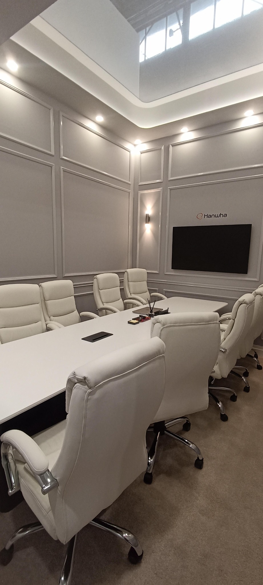 Sala konferencyjna z białym stołem i krzesłami, ściany w szarej tonacji z dekoracyjnymi listwami. Na ścianie logo firmy Hanwha i duży ekran.