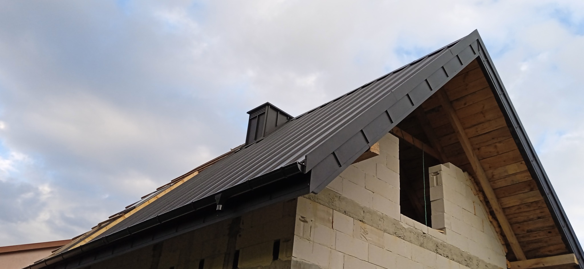 Nowy, grafitowy dach z blachy trapezowej na budynku w trakcie budowy, widok z dołu. Widoczne rynny i komin. W tle pochmurne niebo.