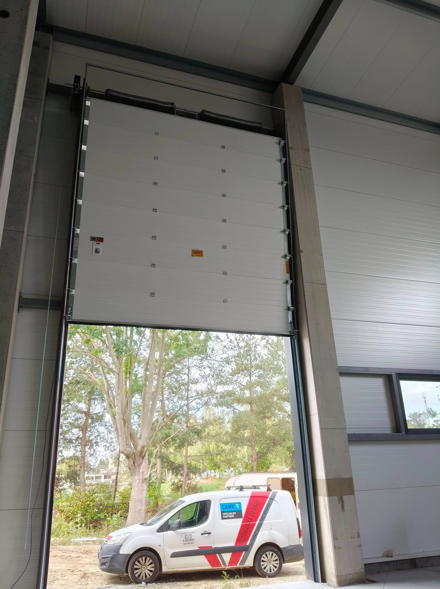 Otwarta segmentowa brama garażowa w hali przemysłowej, z widokiem na zewnątrz i zaparkowany samochód serwisowy z logo firmy. Widoczne sprężyny i mechanizm bramy.