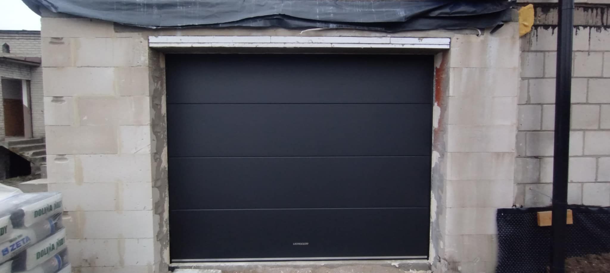 Ciemnoszara segmentowa brama garażowa w surowym otworze budynku z betonu komórkowego. Widoczne worki z materiałami budowlanymi i czarna rura spustowa.