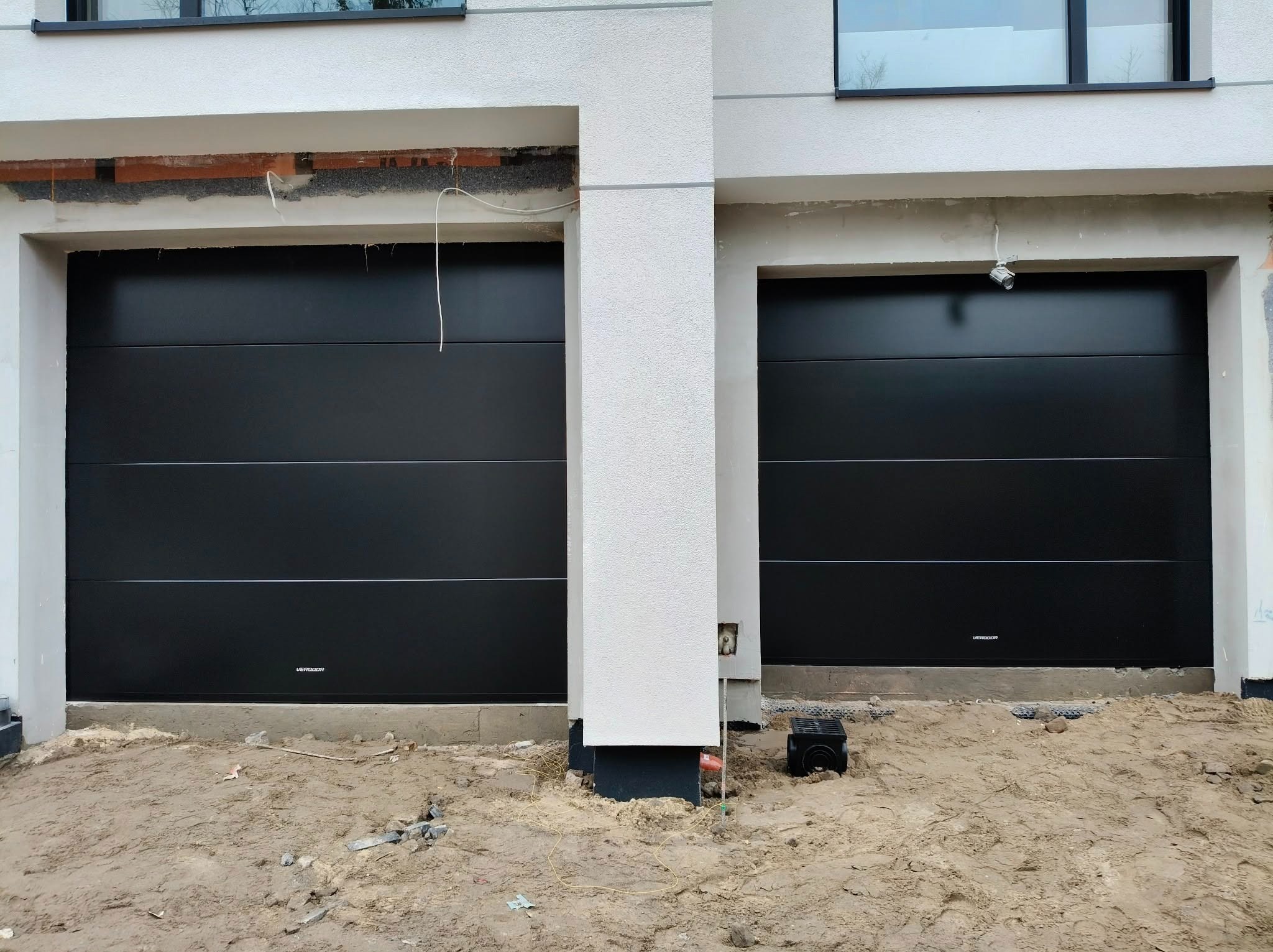 Dwie czarne, segmentowe bramy garażowe w nowoczesnym domu z białymi ścianami. Widoczne niedokończone prace wykończeniowe wokół bram i na podłożu.