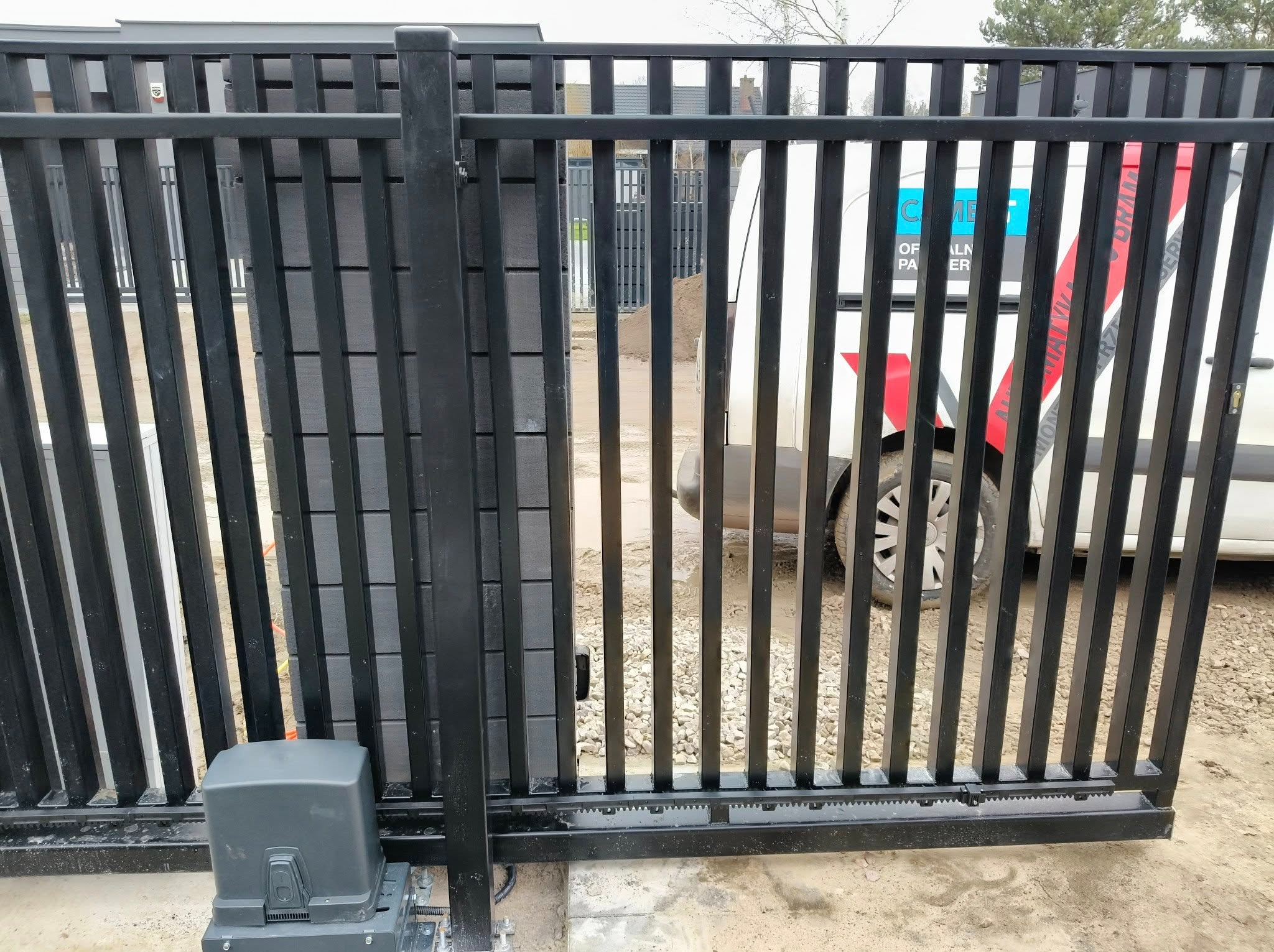 Czarna, przesuwna brama wjazdowa z automatyką, widoczny napęd bramy. W tle zaparkowany samochód dostawczy z reklamą firmy.