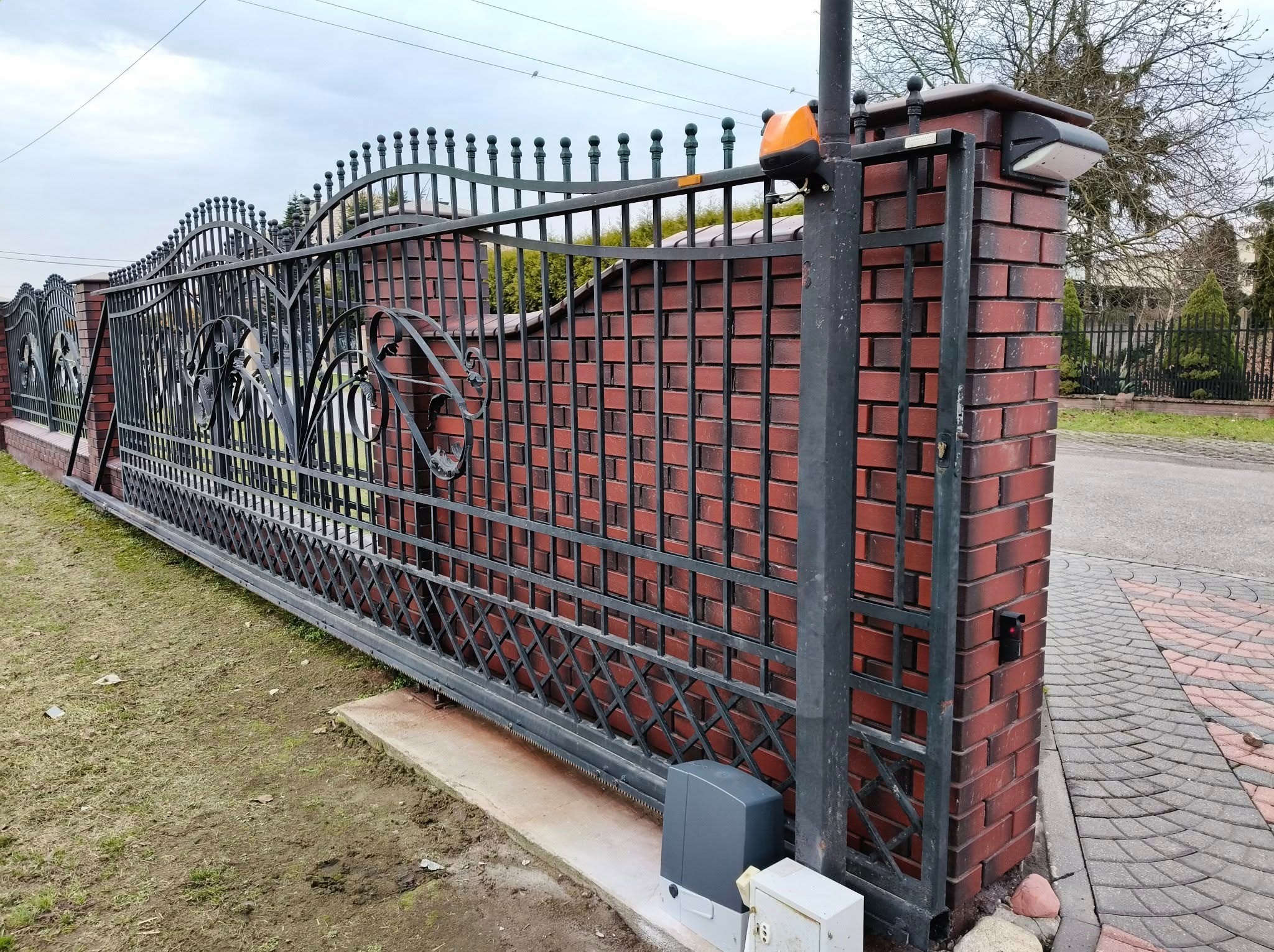 Przesuwna brama wjazdowa z czarnej stali, zdobiona ornamentami, zamontowana na murowanym, czerwonym słupku z cegły klinkierowej. Widoczny napęd i lampa ostrzegawcza.