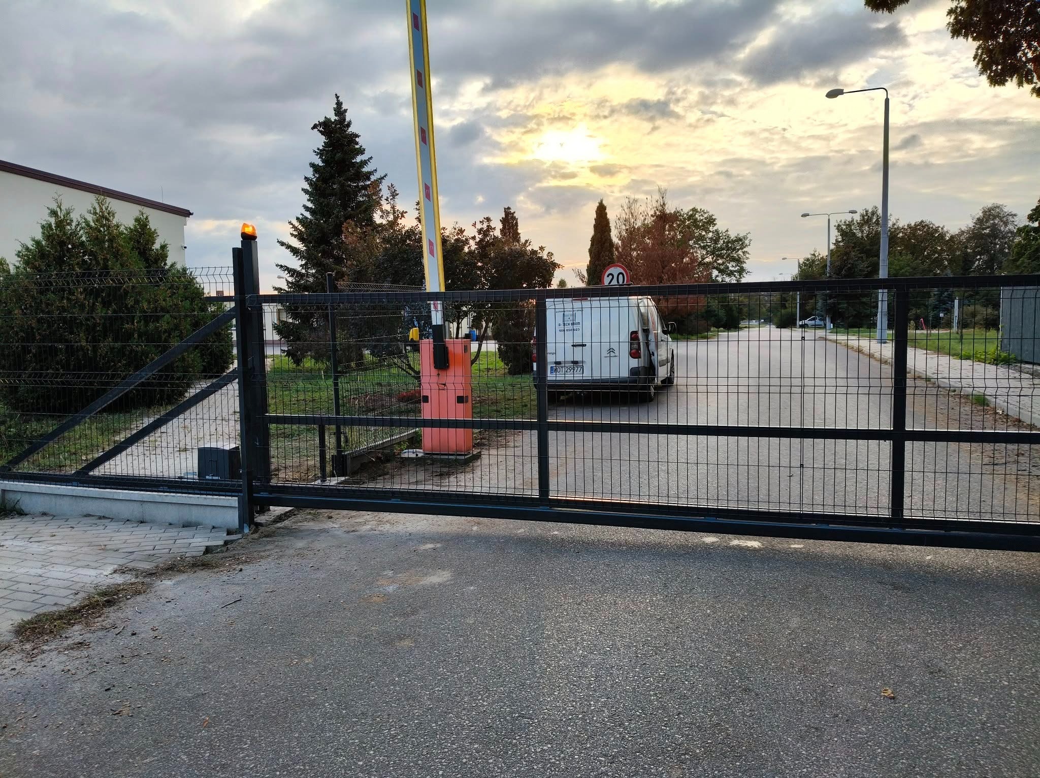 Automatyczna brama wjazdowa z czarnej siatki, z pomarańczowym sygnalizatorem i szlabanem. W tle budynek i biały samochód dostawczy.