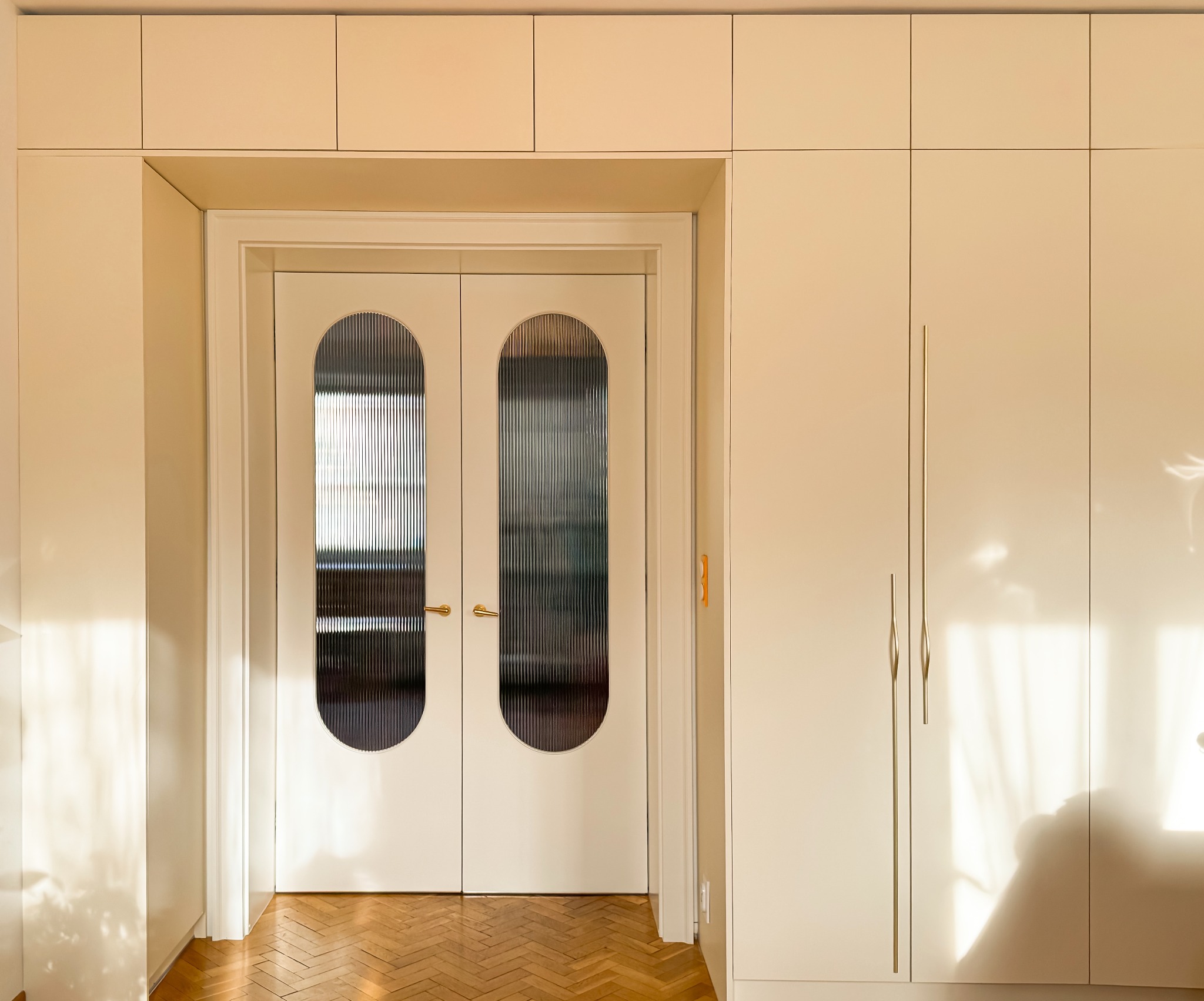 Beżowe, dwuskrzydłowe drzwi wewnętrzne z ryflowanym szkłem w owalnych przeszkleniach, złote klamki, parkiet w jodełkę, zabudowa meblowa w kolorze drzwi.