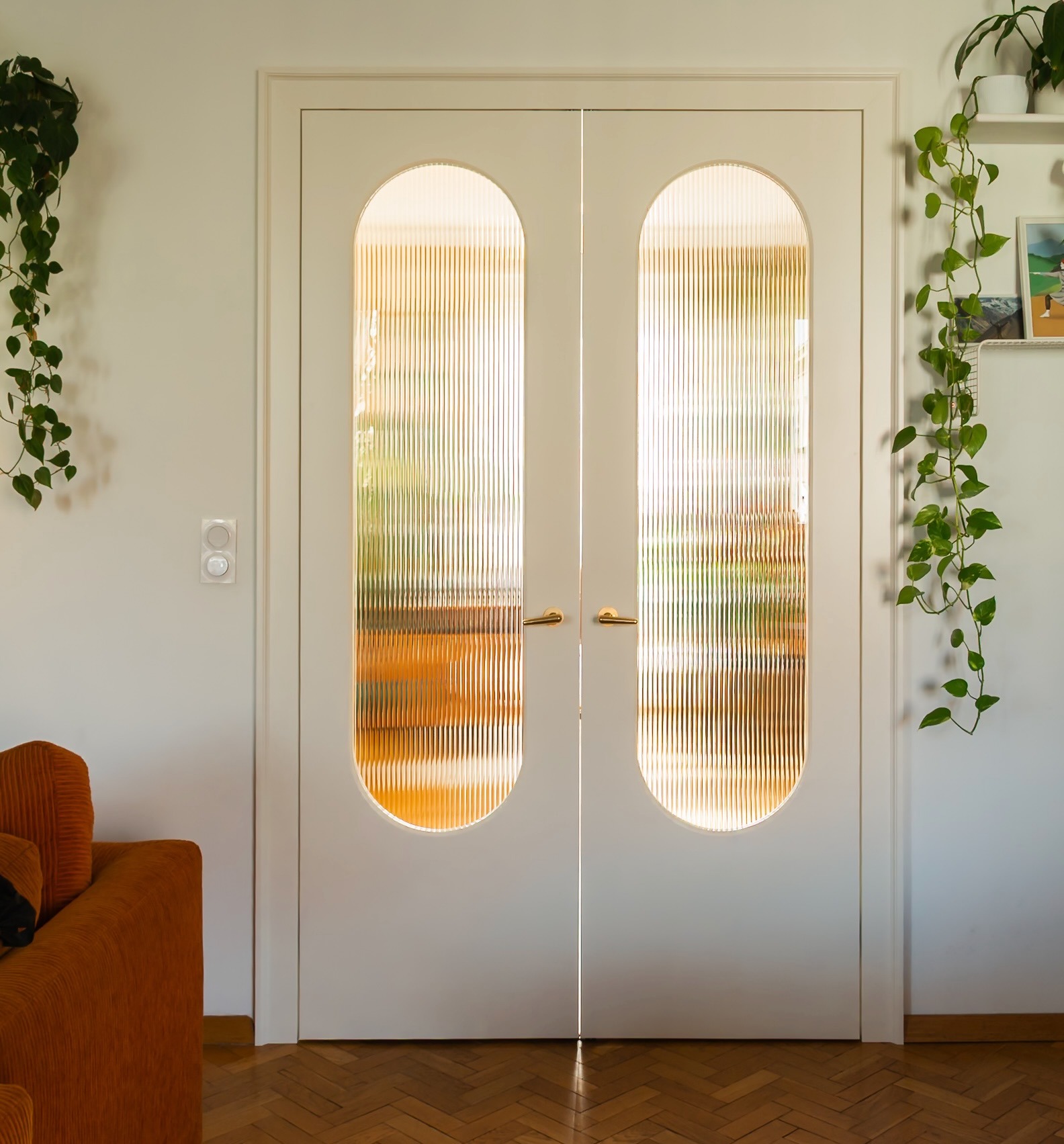 Białe, dwuskrzydłowe drzwi wewnętrzne z ryflowanymi, owalnymi przeszkleniami, złote klamki. W tle widoczny fragment pomarańczowej sofy i parkiet w jodełkę.
