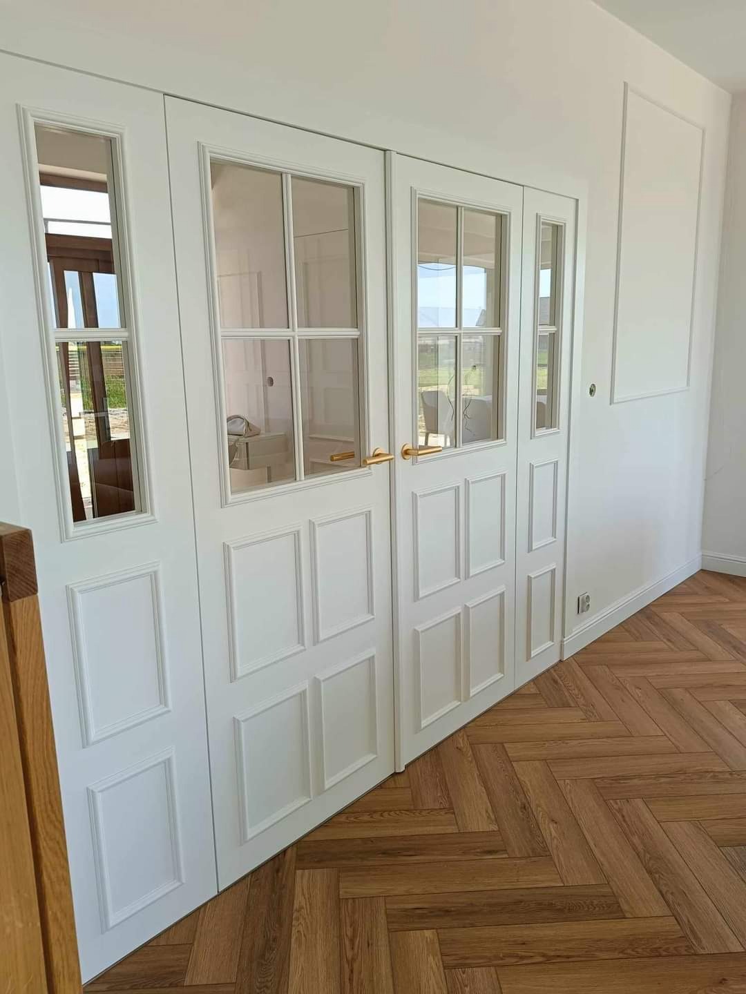 Białe, przeszklone drzwi dwuskrzydłowe z ozdobnymi panelami, złote klamki, parkiet w jodełkę, subtelne oświetlenie, eleganckie wykończenie wnętrza.
