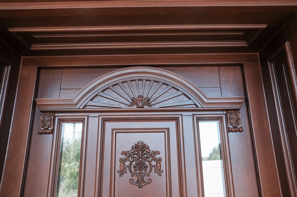 Fragment bogato zdobionych, ciemnobrązowych drzwi z przeszkleniami i dekoracyjnymi elementami rzeźbionymi w drewnie, widoczne od góry.