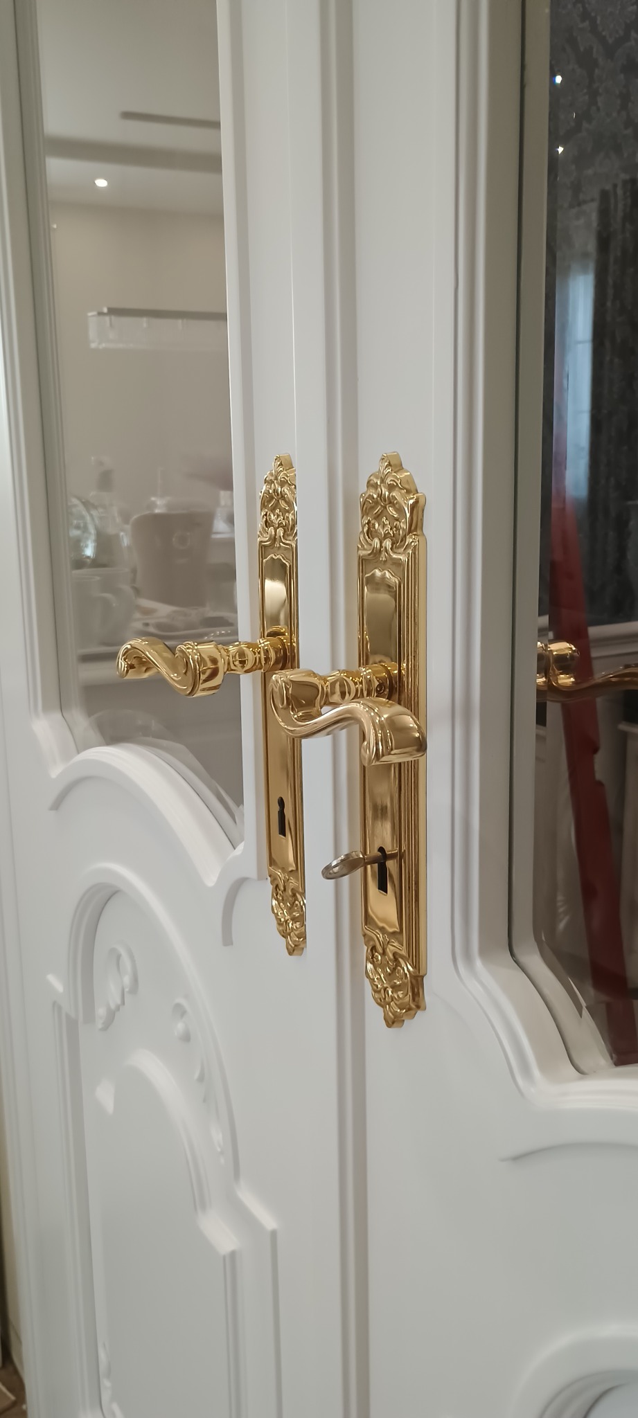 Białe, przeszklone drzwi z ozdobnymi, złotymi klamkami i kluczem w zamku. Widoczne detale frezowania na skrzydle drzwiowym i odbicie wnętrza w szybie.