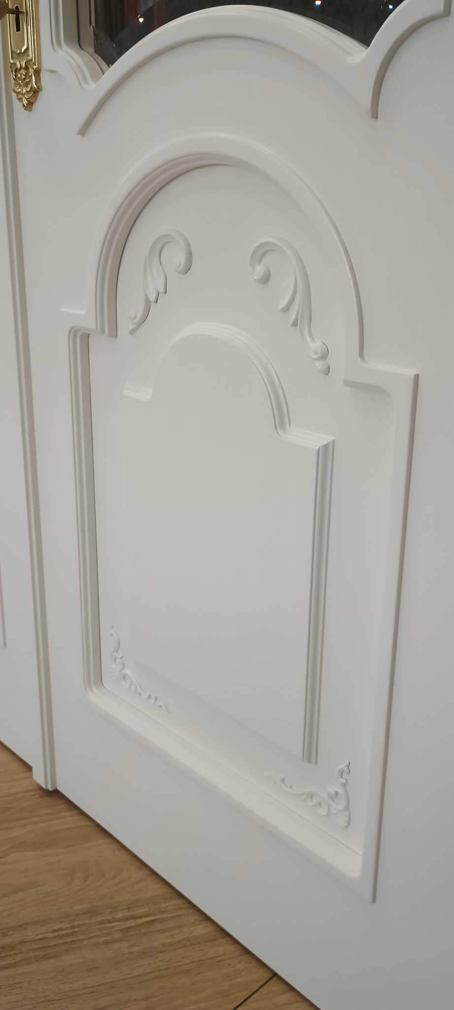 Białe drzwi wewnętrzne z dekoracyjnymi frezami i złotą klamką, widoczne pod kątem. Klasyczny design z subtelnymi zdobieniami.