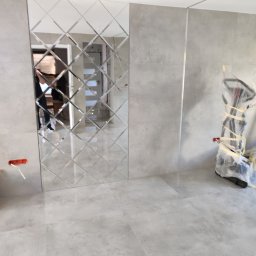 EXPERT Ryszard Studniarz - Ściana z dekoracyjnych, lustrzanych płytek w kształcie rombów, odbijających wnętrze. Szare płytki podłogowe i fragment sprzętu zabezpieczonego folią.