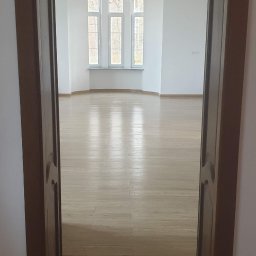 EXPERT Ryszard Studniarz - Jasne, przestronne wnętrze z drewnianą podłogą, widoczne przez otwarte, ciemne drzwi. Trzy okna w głębi wpuszczają naturalne światło. Minimalistyczny wystrój.