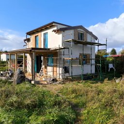 EXPERT Ryszard Studniarz - Dom w trakcie ocieplania elewacji, widoczne rusztowania, okna zabezpieczone folią, częściowo otynkowana fasada, prace w toku, trawa i zieleń wokół budynku.