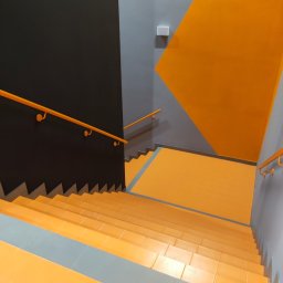 EXPERT Ryszard Studniarz - Schody z pomarańczowymi stopniami i szarą wstawką, czarna ściana z pomarańczową poręczą, szara ściana z pomarańczowym trójkątem. Widok z góry.