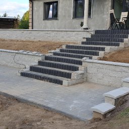 EXPERT Ryszard Studniarz - Wykończony taras z kostki brukowej i schodami z bloków betonowych, otoczony ziemią. Widok na budynek z oknami i meblami ogrodowymi.
