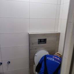 EXPERT Ryszard Studniarz - Montaż toalety w wykończonej łazience z białymi płytkami i drewnopodobnym akcentem. Widoczny instalator w firmowej koszulce podczas pracy.