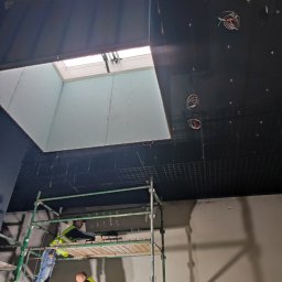 EXPERT Ryszard Studniarz - Montaż sufitu podwieszanego z płyt kartonowo-gipsowych w biurze. Dwóch pracowników na rusztowaniu, widoczne kable i kratownica sufitowa. Otwór okienny w suficie.