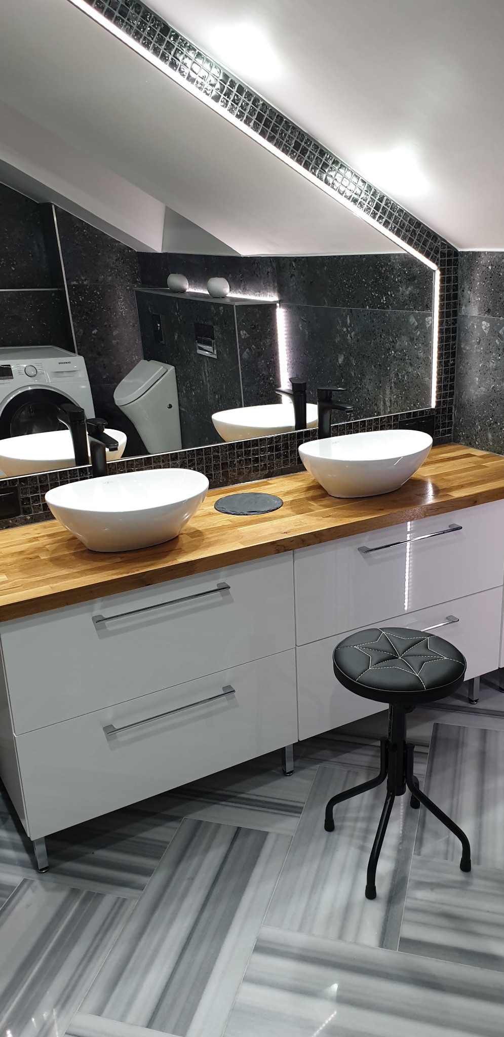 Nowoczesna łazienka z dwoma białymi umywalkami na drewnianym blacie, białe szafki z chromowanymi uchwytami, marmurowa podłoga i czarne płytki na ścianie.