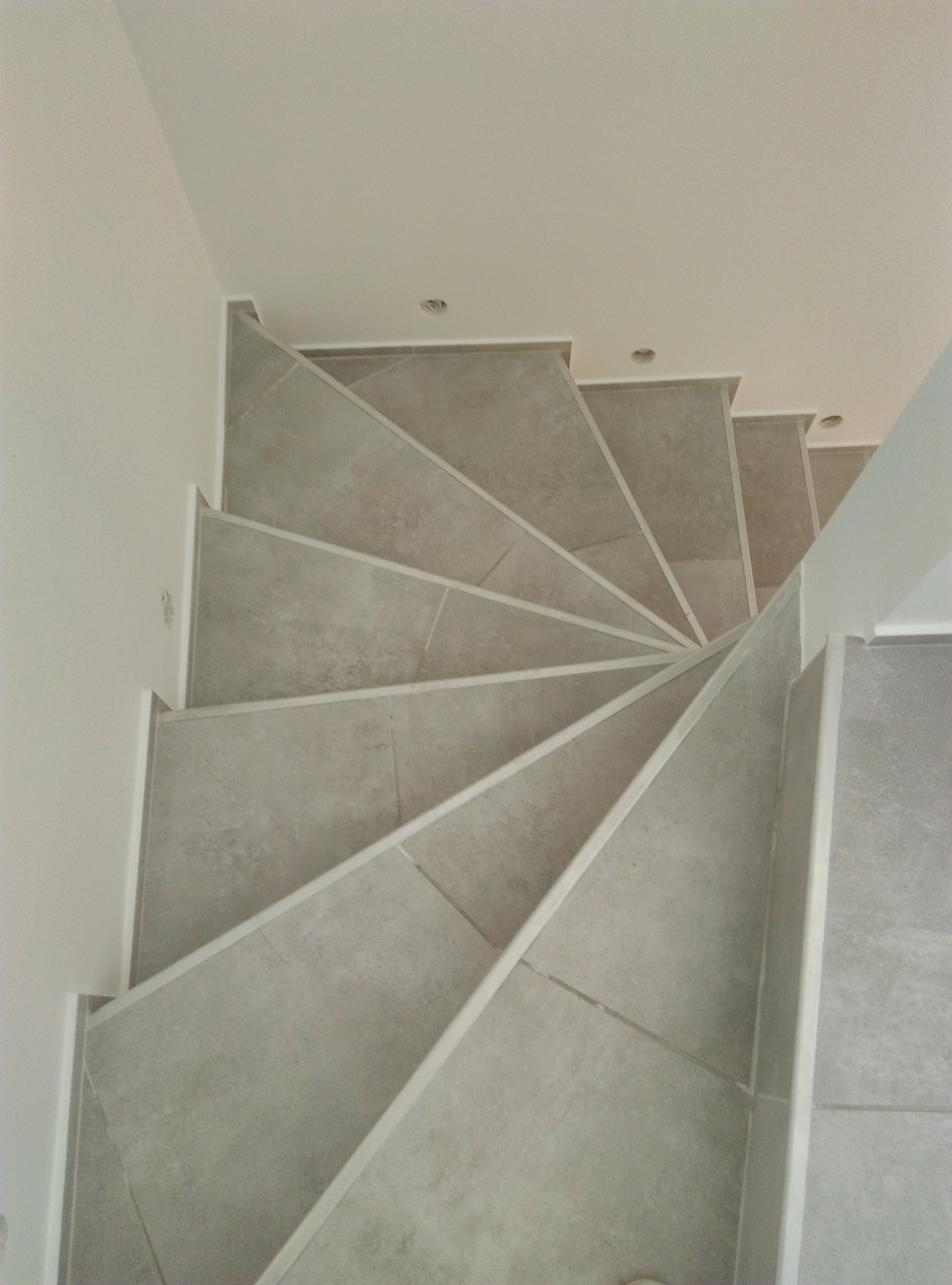 Spiralne schody wyłożone szarymi płytkami z białymi listwami, widok z góry, oświetlenie punktowe w suficie, minimalistyczny design.