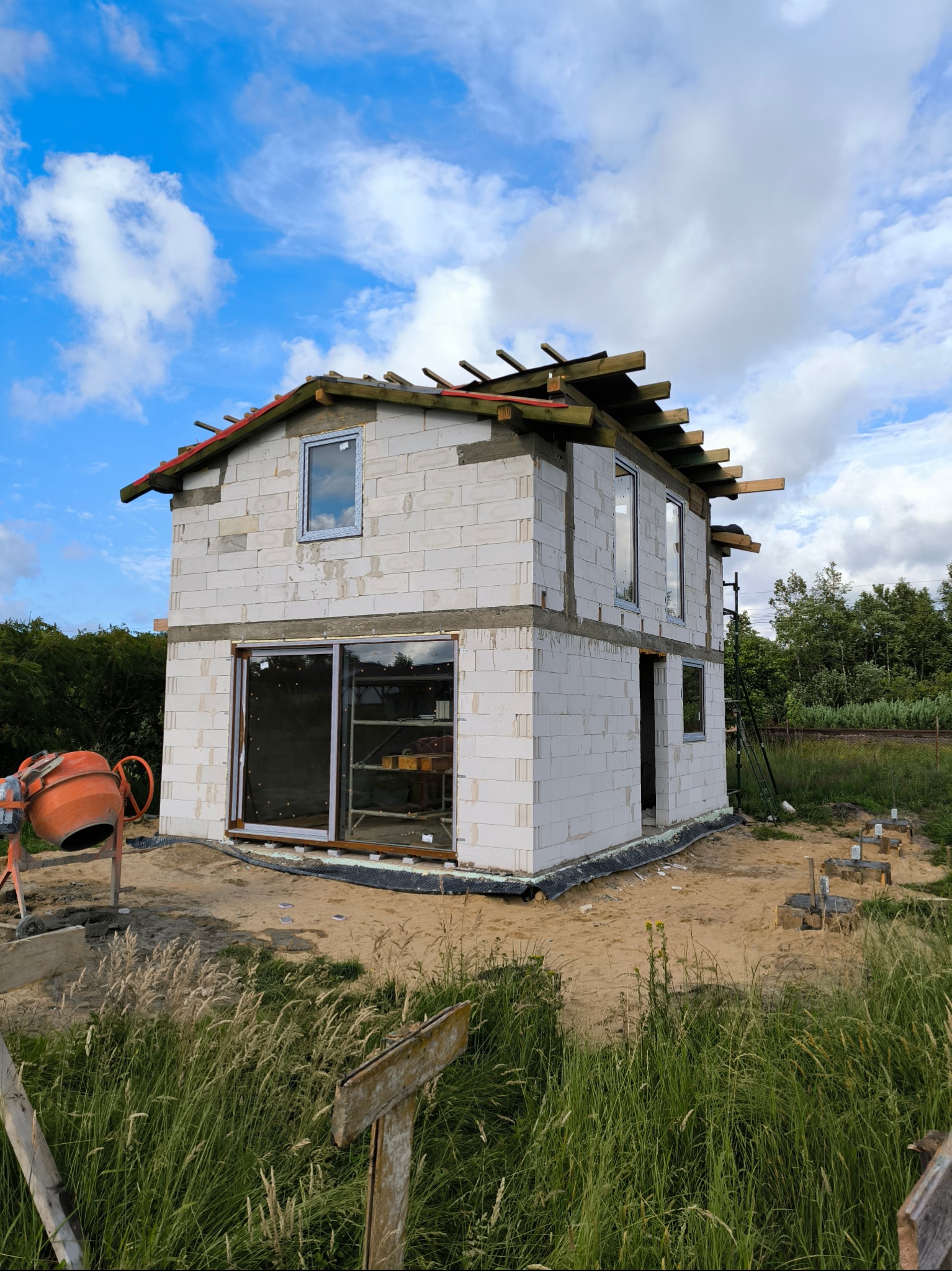 Budowa domu z betonu komórkowego w trakcie realizacji, widoczne okna, pustaki, elementy konstrukcji dachu i betoniarka. Fundamenty w tle, błękitne niebo.