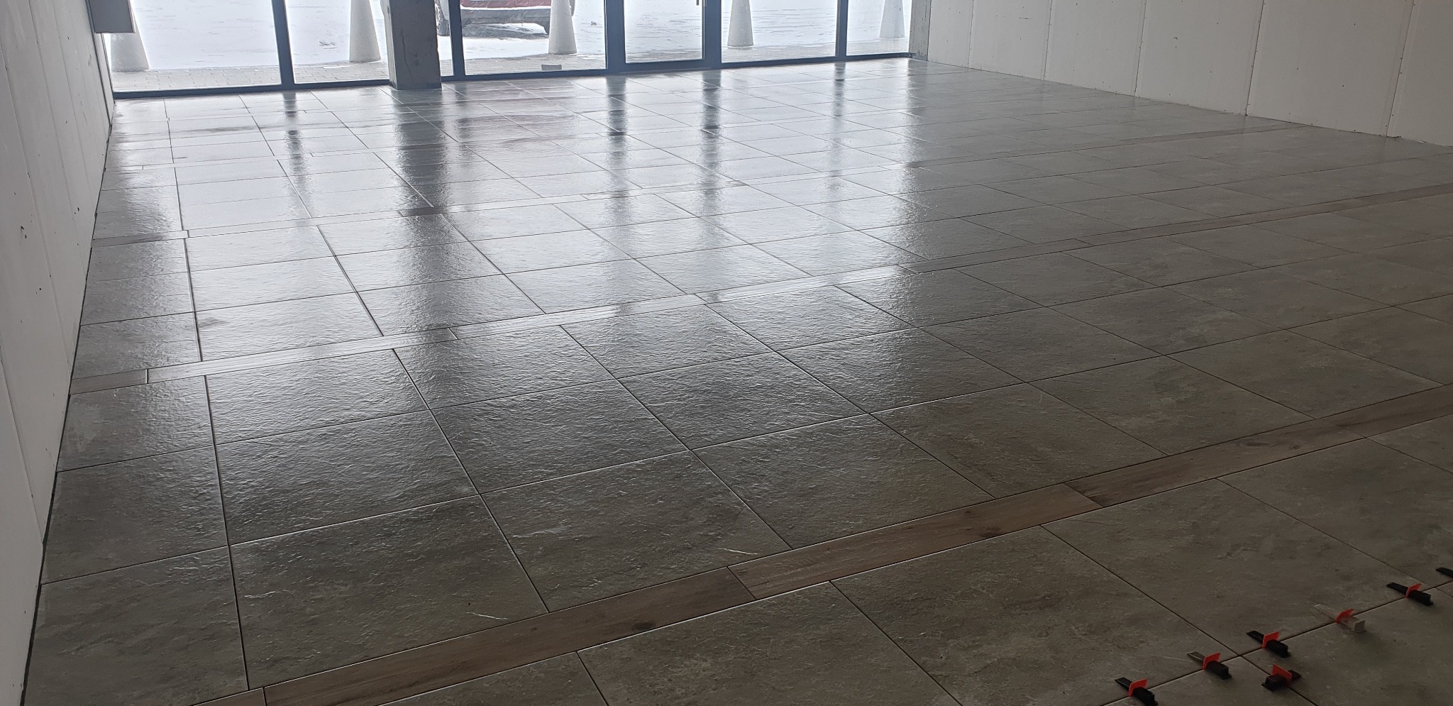 Ułożona podłoga z płytek w odcieniach szarości z drewnianymi wstawkami, widoczne krzyżyki dystansowe. Duże okna z widokiem na zewnątrz.