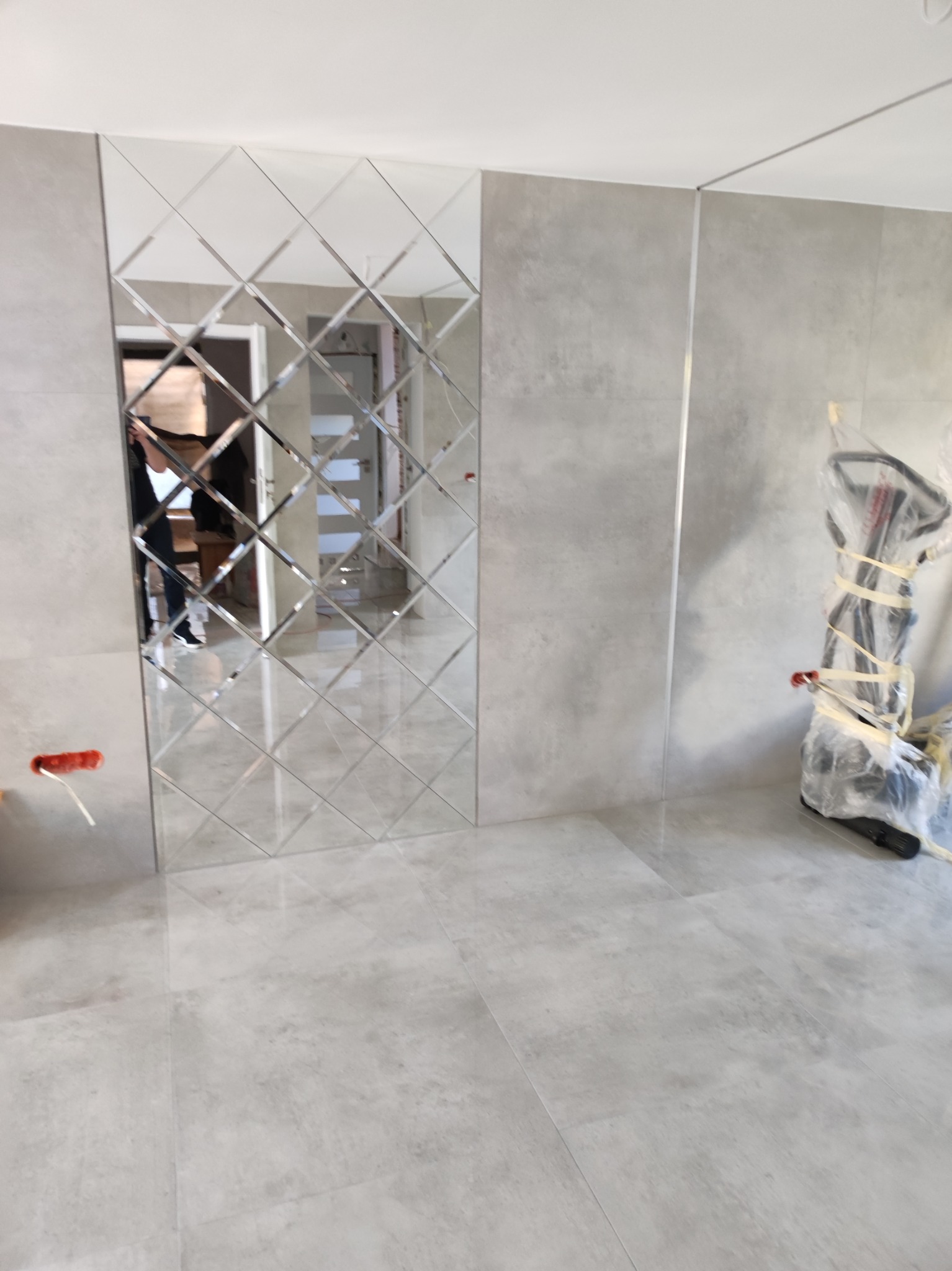 Ściana z dekoracyjnych, lustrzanych płytek w kształcie rombów, odbijających wnętrze. Szare płytki podłogowe i fragment sprzętu zabezpieczonego folią.