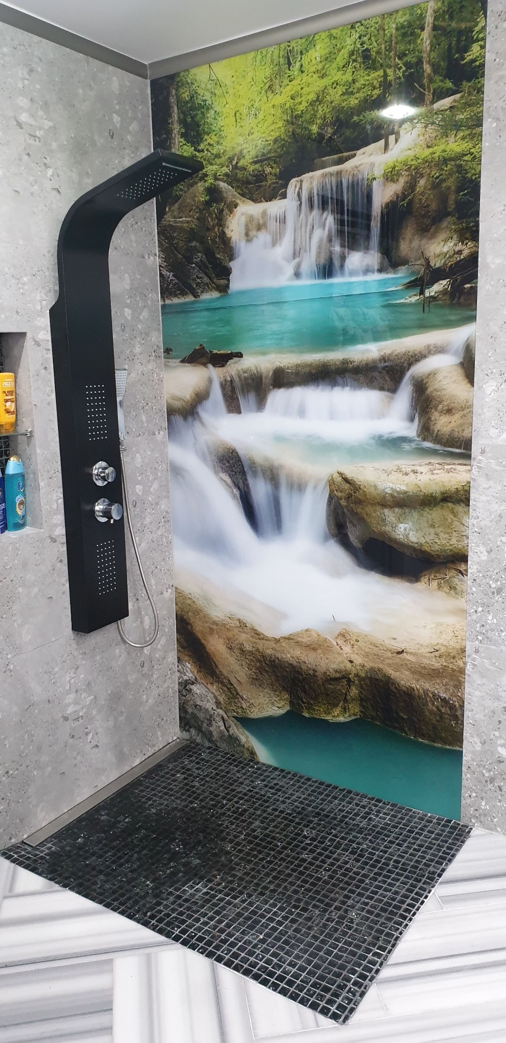 Nowoczesna kabina prysznicowa z panelem wodospad w Maszewie Lęborskim. Czarny panel natryskowy na tle szarej ściany i fototapety z wodospadem. Ciemna mozaika na podłodze.