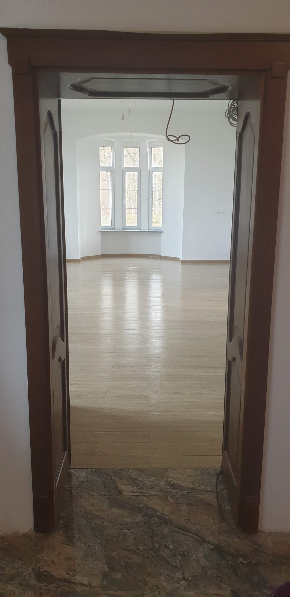 Jasne, przestronne wnętrze z drewnianą podłogą, widoczne przez otwarte, ciemne drzwi. Trzy okna w głębi wpuszczają naturalne światło. Minimalistyczny wystrój.