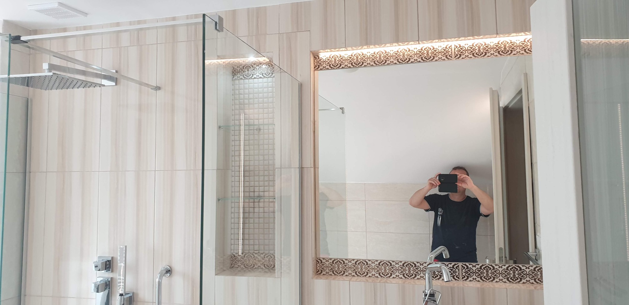 Wykończona łazienka z kabiną prysznicową typu walk-in, lustrem z dekoracyjnym oświetleniem i wnęką z mozaiką. Osoba robiąca zdjęcie widoczna w lustrze.