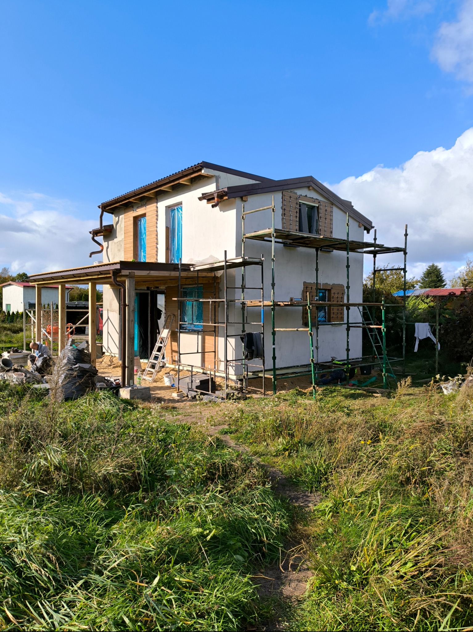 Dom w trakcie ocieplania elewacji, widoczne rusztowania, okna zabezpieczone folią, częściowo otynkowana fasada, prace w toku, trawa i zieleń wokół budynku.