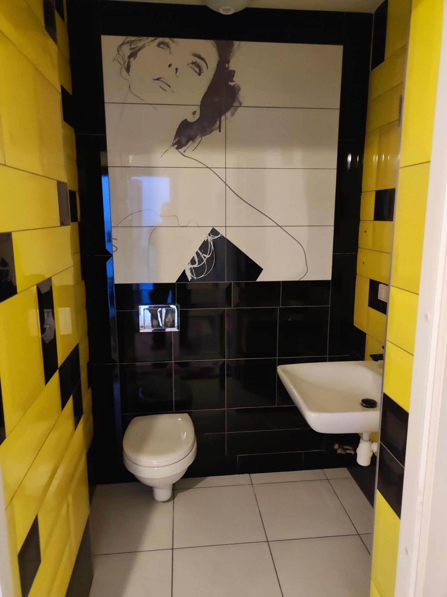 Nowoczesna łazienka z czarno-białymi płytkami i żółtymi akcentami. Na ścianie grafika kobiety. Umywalka i toaleta w kolorze białym. Minimalistyczny design.