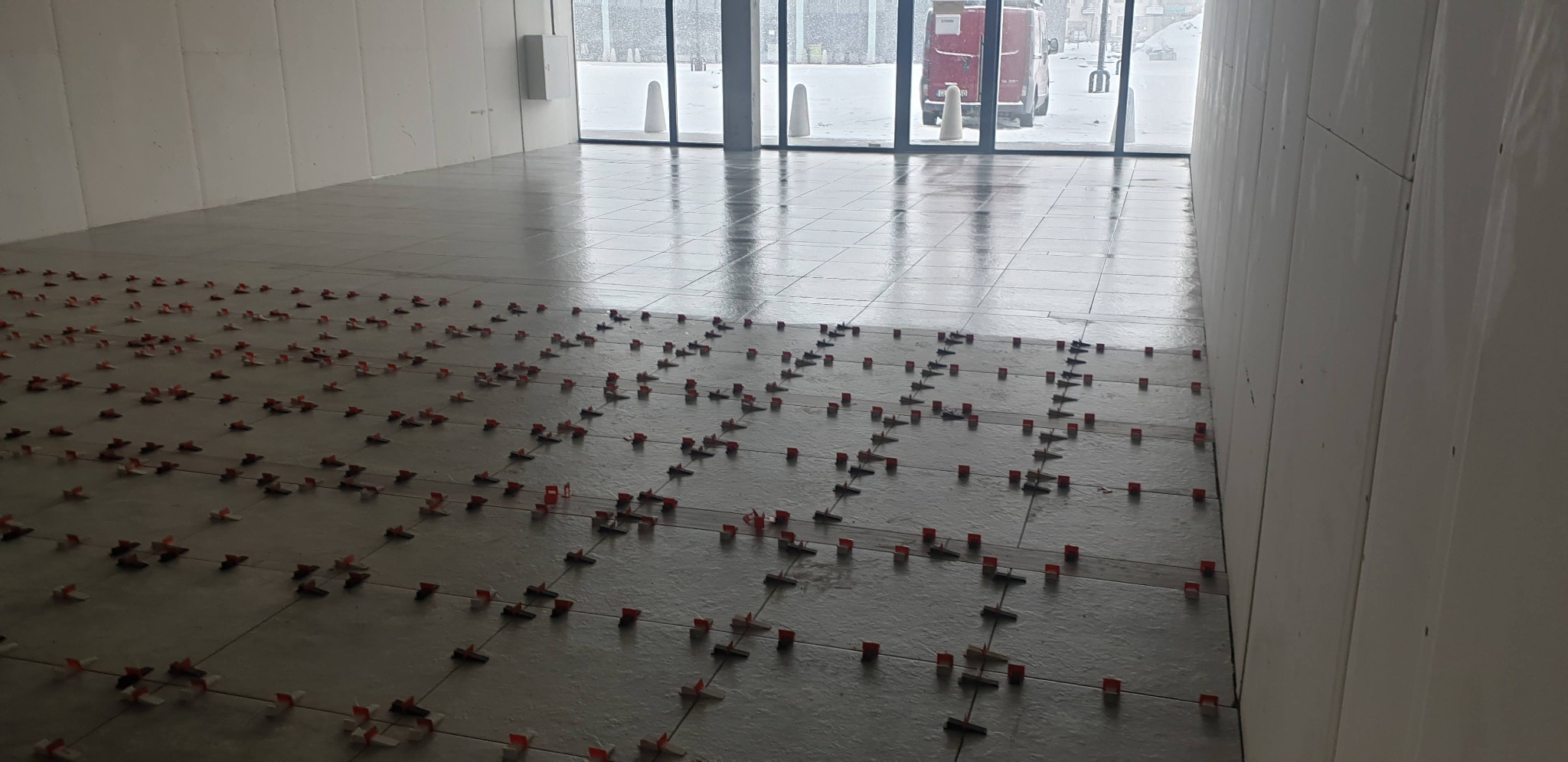 Układanie szarych płytek podłogowych z użyciem czerwonych klipsów poziomujących w jasnym, przestronnym wnętrzu z dużymi oknami i ścianami z płyt kartonowo-gipsowych.