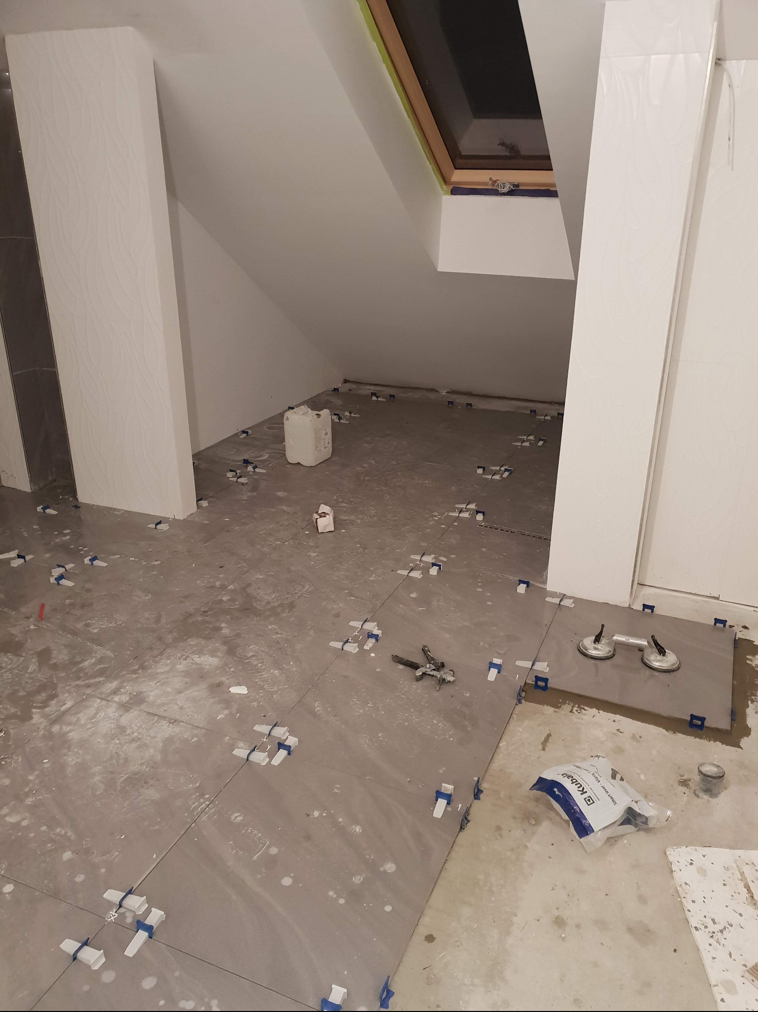 Układanie szarych płytek podłogowych w skosach poddasza z oknem dachowym. Widoczne narzędzia glazurnicze i system poziomowania płytek.