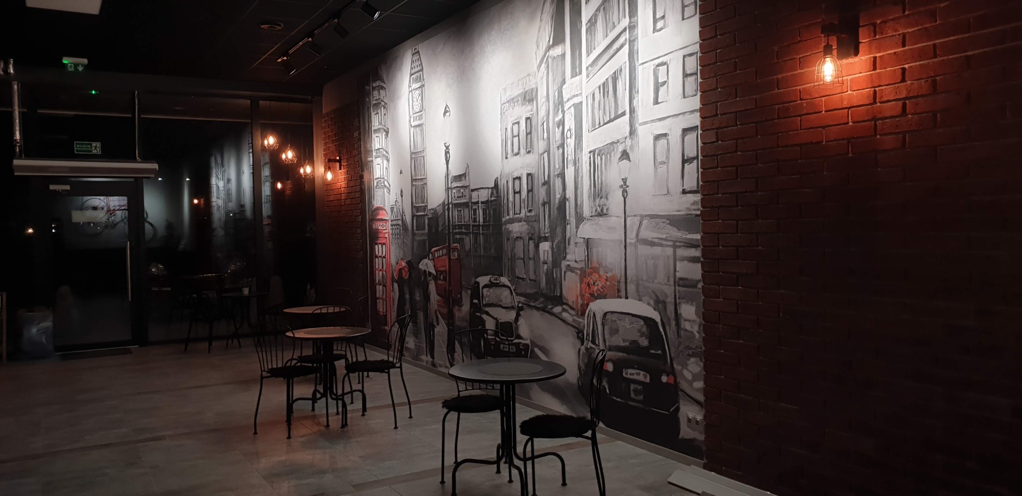 Wnętrze biura z muralem Londynu na ścianie i ceglaną ścianą z oświetleniem. Widoczne stoliki z krzesłami, tworzące przestrzeń do spotkań.