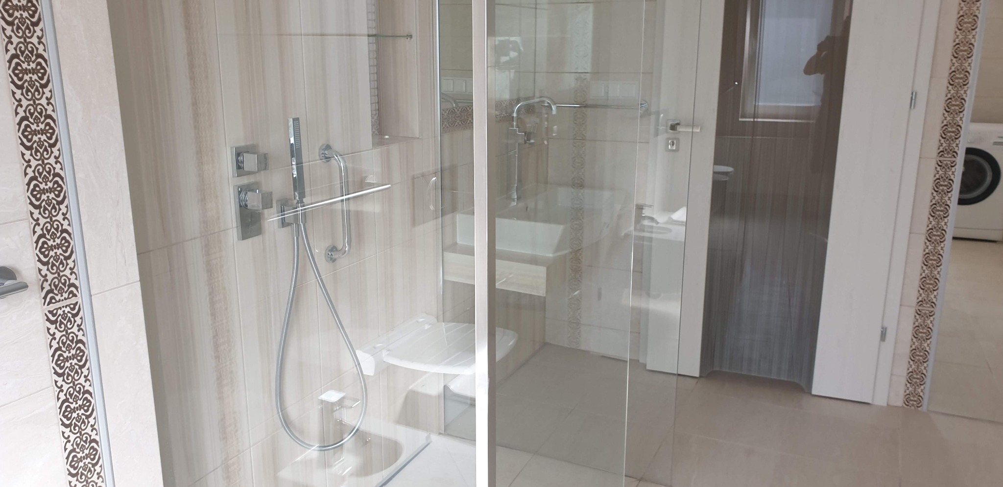 Nowoczesna kabina prysznicowa z siedziskiem i armaturą w odcieniach beżu, wykończona dekoracyjnym pasem płytek. Widoczna umywalka i pralka w tle.