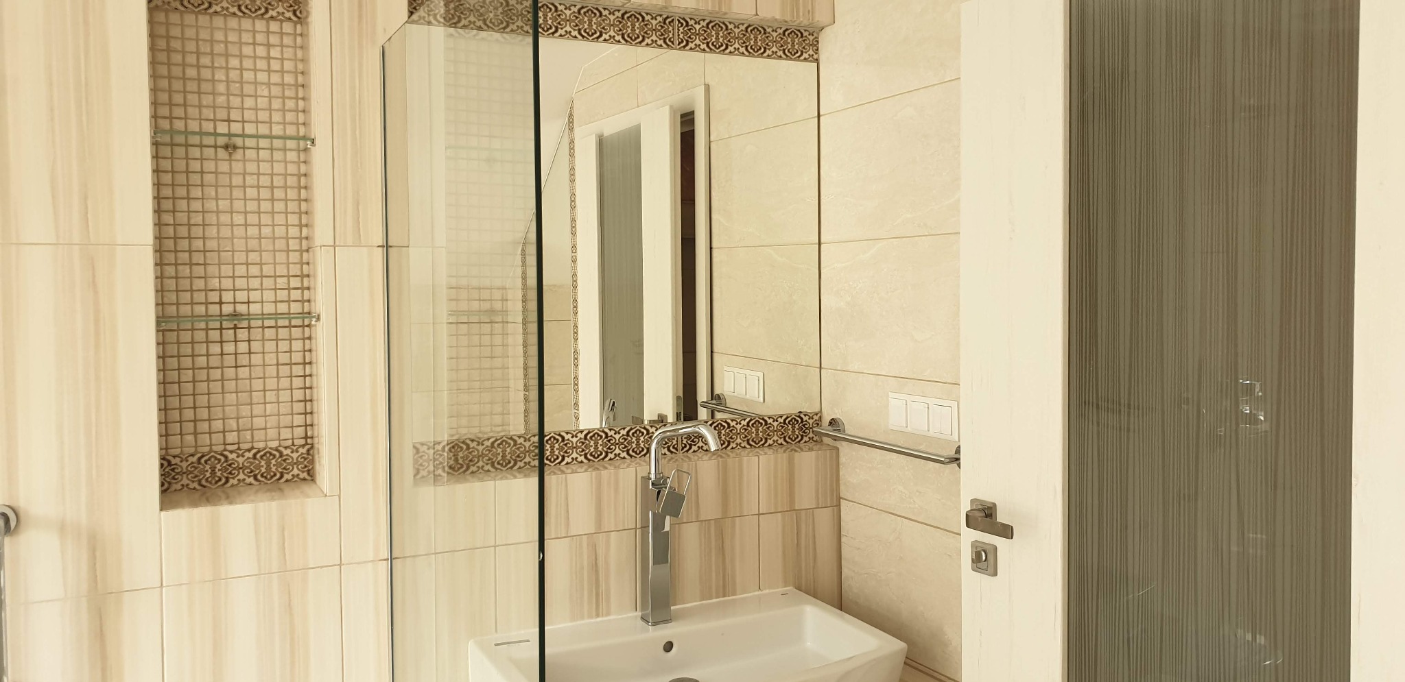 Beżowa łazienka z umywalką, lustrem z dekoracyjnym obramowaniem i wnęką z mozaiką. Widoczna armatura i fragment drzwi.