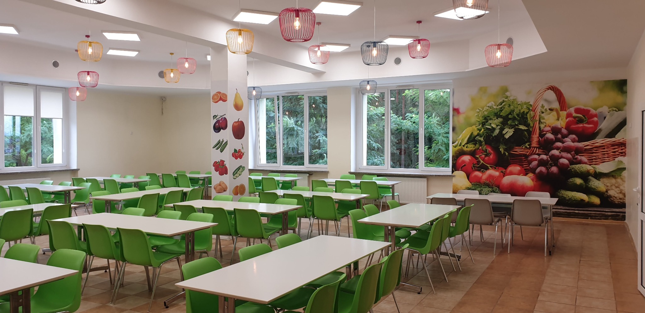 Jasna stołówka z zielonymi krzesłami i białymi stołami, ozdobiona kolorowymi lampami i fototapetą z motywem owoców i warzyw. Widok na zieleń za oknem.