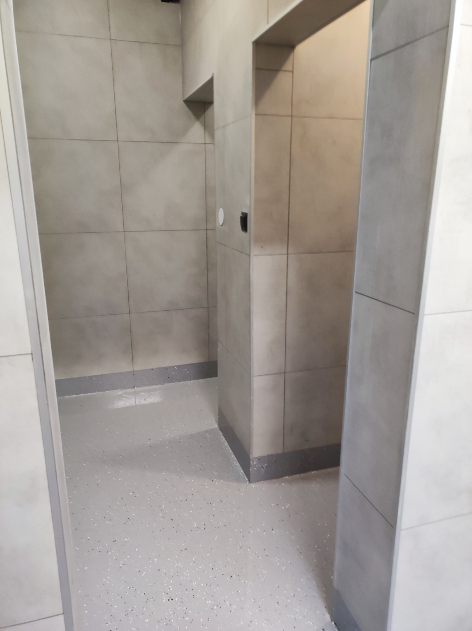 Wykończona łazienka z szarymi, kwadratowymi płytkami na ścianach i podłodze z żywicy epoksydowej z brokatem. Widoczne detale wykończeniowe i oświetlenie.