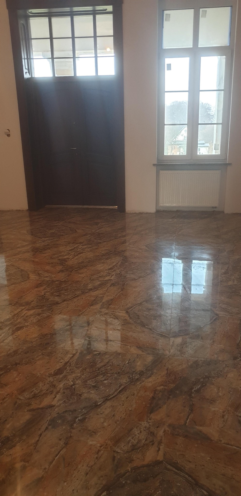Błyszcząca, brązowa podłoga z płytek imitujących marmur w nowym wnętrzu. Widoczne okno i drzwi z szybami. Świeżo położona podłoga po remoncie.