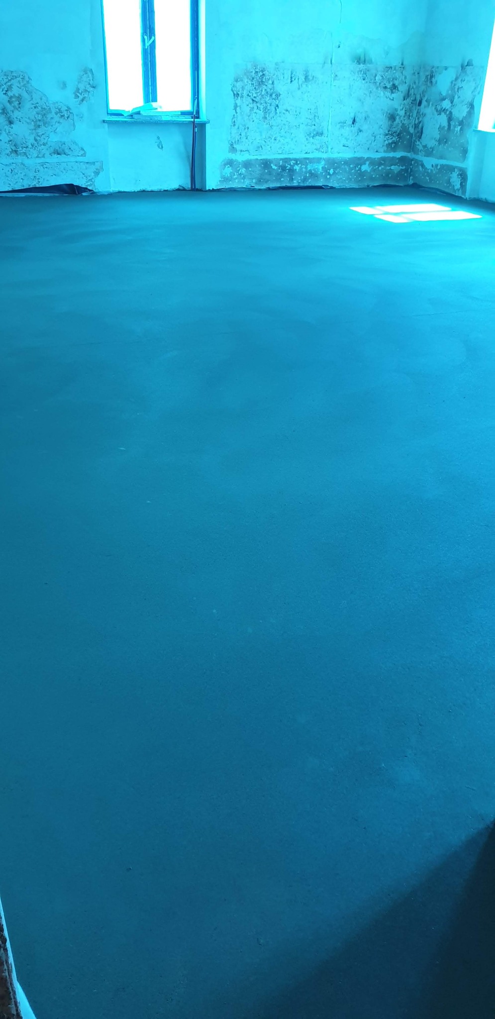 Wylana, gładka, turkusowa posadzka w pomieszczeniu z oknem i fragmentami starego tynku na ścianach. Widoczne ślady wcześniejszych prac remontowych.