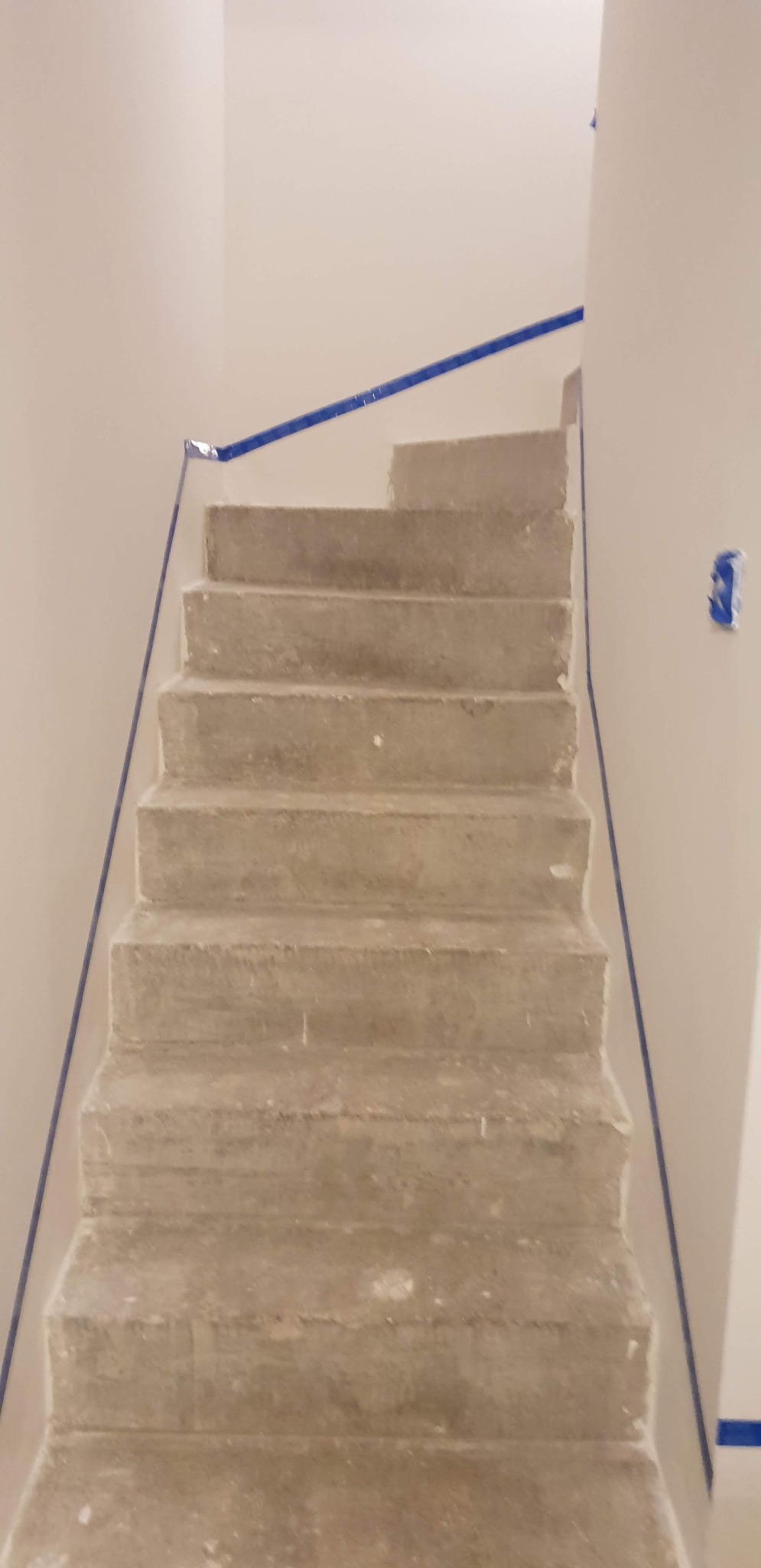 Surowe betonowe schody w trakcie wykańczania, zabezpieczone niebieską taśmą malarską na ścianach i stopniach. Widoczne niedoskonałości betonu i przygotowanie do dalszych prac.