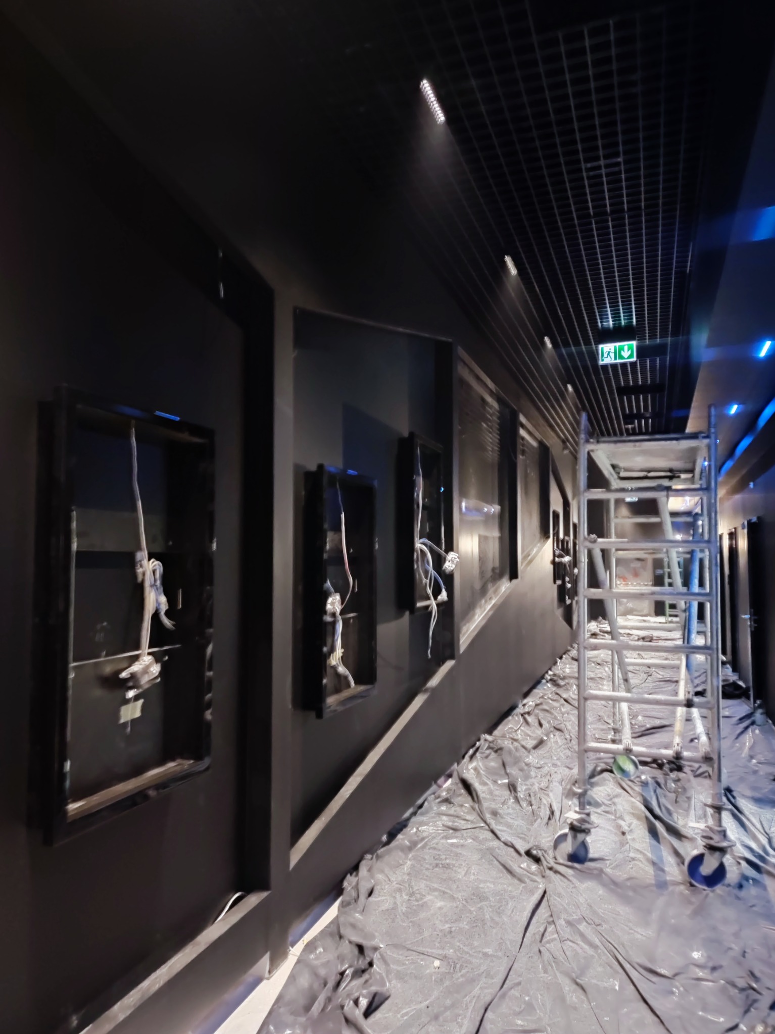 Ciemne wnętrze w trakcie prac wykończeniowych: ściany z otworami na instalacje, zabezpieczona podłoga folią i aluminiowe rusztowanie. Widoczna kratownica sufitu.