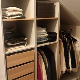 Firma Budowlana Piotr Milewski - Biała szafa wnękowa z drewnianymi szufladami i półkami, wypełniona ubraniami i koszem, dopasowana do skosu dachu, na drewnianej podłodze.