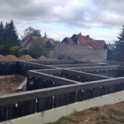 Firma Budowlana Piotr Milewski - Zabezpieczone fundamenty domu jednorodzinnego z izolacją pionową i poziomą, widoczne zbrojenie. W tle zabudowa jednorodzinna i drzewa.