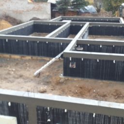 Firma Budowlana Piotr Milewski - Zabezpieczone fundamenty domu jednorodzinnego z izolacją pionową i szalunkiem traconym, widok z góry na plac budowy. W tle hałdy piasku i zabudowa.