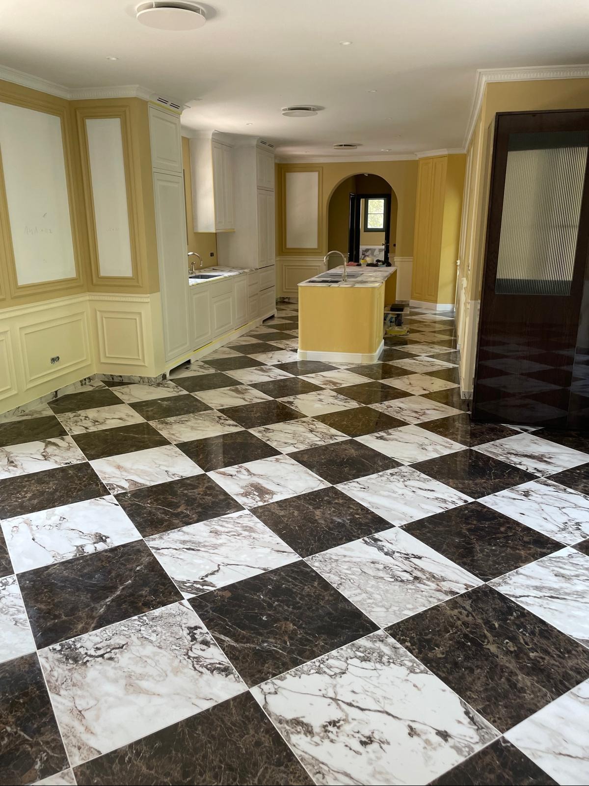 Kuchenne wnętrze z błyszczącą, szachownicową podłogą z płytek imitujących marmur. W tle żółte ściany i białe szafki kuchenne. Widoczny fragment wyspy kuchennej.