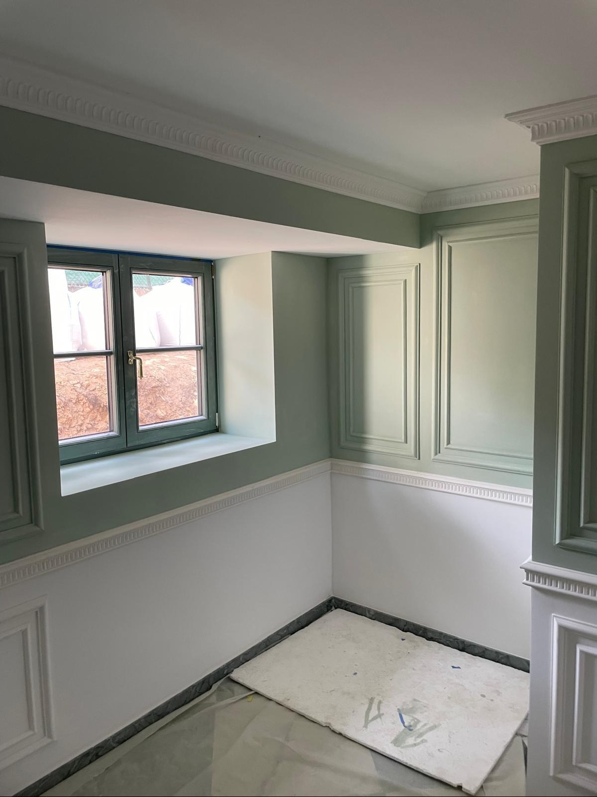 Ściana po malowaniu w odcieniach zieleni z dekoracyjnymi panelami i listwami. Widoczne okno z zieloną ramą i zabezpieczona podłoga. Klasyczny styl wykończenia.